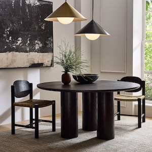 Könnte beinhalten: Ein runder Esstisch aus dunklem Holz mit drei zylindrischen Beinen, umgeben von zwei schwarzen Stühlen mit gewebten Sitzen. Über dem Tisch hängen zwei Pendelleuchten, und eine Vase mit Grün steht auf dem Tisch. Ein großes abstraktes Gemälde hängt an der Wand.