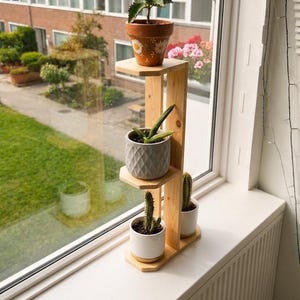 May include: A tiered wooden plant stand with three levels holding four potted plants. The top level holds a terracotta pot with white daisy designs. The middle level holds a grey textured pot. The bottom level holds two white pots.