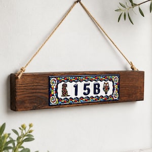 May include: A rustic wooden address plaque with a decorative ceramic tile. The tile features a dog and an owl flanking the number 15B, set against a blue background with colorful accents. The plaque is suspended by a natural rope.