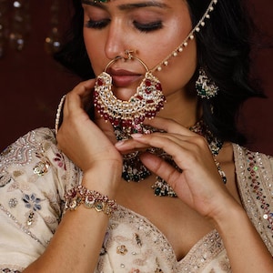 May include: A large gold-toned nose ring featuring red and clear stones with dangling beads, connected by a delicate chain to a matching earring. The jewellery set includes a coordinating bracelet and necklace, all adorned with intricate stone work.