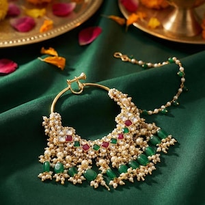 Puede incluir: Un intrincado aro de nariz dorado con un aro grande y una cadena colgante. La joya está adornada con perlas blancas, piedras preciosas de color verde esmeralda y rojo rubí. El fondo es de tela de satén verde oscuro.