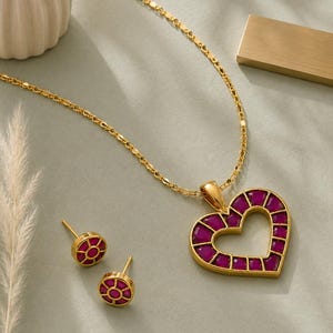 Puede incluir: Un collar de tono dorado con un colgante en forma de corazón incrustado con pequeñas piedras magenta de corte cuadrado, acompañado de un par de pendientes a juego. Cada pendiente consiste en un marco circular dorado con una piedra central.