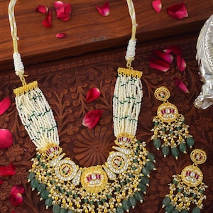 May include: An ornate gold necklace and matching earrings set. The necklace features a central pendant with red and green accents, surrounded by white beads and green gemstone drops. The earrings mirror the necklace design. The set is displayed on a wooden surface.