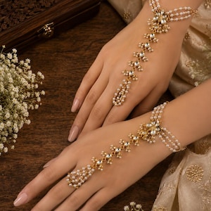 May include: A gold-toned hand harness featuring three delicate strands of small white pearls connected to a central floral motif. The design includes a chain extending to a pearl-encrusted ring loop, accented with clear stones throughout the structure.