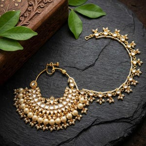 May include: Gold-coloured nose ring with a pearl and crystal design. The jewellery features a large, ornate crescent shape with dangling pearls and a delicate chain. The piece is set against a dark background, with a wooden box and green leaves in the background.