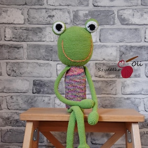 May include: A handmade green crochet frog doll with large black and white eyes, a smiling orange mouth, and long, slender limbs. The frog is seated on a wooden stool. The frog's body is a mix of colorful yarn. The text "Szydełko Oli" is visible.