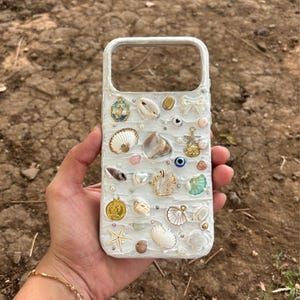 May include: White phone case adorned with seashells, pearls, and small charms. The case features various shells, including scallop and conch shells, with gold-coloured accents and small crystals. The design has a textured, raised surface.