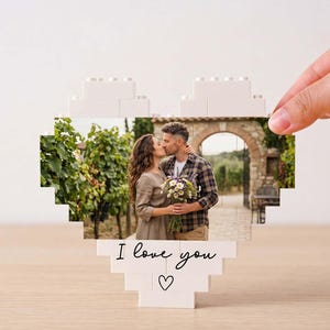 Könnte beinhalten: Ein herzförmiges Display aus ineinandergreifenden weißen Kunststoffbausteinen mit einem aufgedruckten Foto eines Paares, das sich in einem Weinberg umarmt. Der untere Teil der weißen Bausteinstruktur zeigt den schwarzen Text I love you und ein Herz.