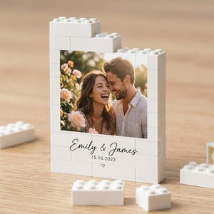 Könnte beinhalten: Ein weißer Bausteinrahmen mit einem rechteckigen Foto eines lächelnden Paares. Der Rahmen enthält den aufgedruckten Text Emily & James sowie das Datum 15.10.2023 mit einem kleinen Herzsymbol. Mehrere lose weiße Bausteine liegen verstreut daneben.