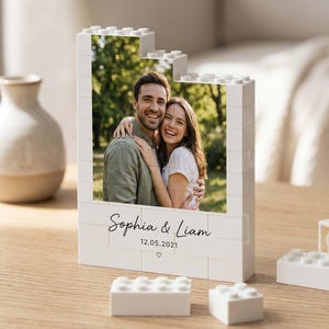 Könnte beinhalten: Ein weißer Bausteinrahmen mit einem aufgedruckten Foto eines lächelnden Paares. Der Rahmen hat eine abgestufte Oberkante und zeigt den Text Sophia & Liam mit dem Datum 12.05.2021 sowie ein kleines Herzsymbol, das in schwarzer Tinte aufgedruckt ist.