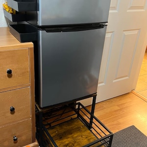 May include: A silver mini-refrigerator with a black top and bottom section, sitting on a black metal frame with a wooden shelf and a pull-out basket. A black shelving unit is to the left. The text "BestdealsArt" is at the top and bottom.