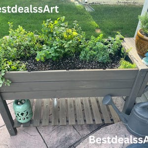May include: A wooden raised garden bed with various green plants, a small ceramic pot, and a decorative figurine. A gray watering can and a green lantern are also visible. The text "BestdealssArt" is displayed.