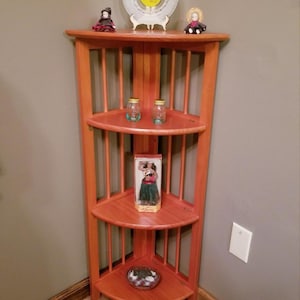 May include: A wooden corner shelf unit with three shelves, holding various decorative items. The shelves are a reddish-brown color, and the unit is positioned in a corner. The text "BestdealssArt" is visible.