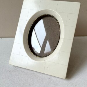 May include: Ivory-colored rectangular picture frame with an oval opening. The frame has a gold-colored inner rim and a textured surface. The frame is angled slightly, showcasing its design and details.