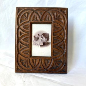 Op de afbeelding: Een vintage houten fotolijst met een gesneden geometrisch ontwerp. De lijst bevat een zwart-witfoto van een vrouw en een mopshond. De lijst is rechthoekig en heeft een donkerbruine afwerking.