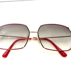 May include: Vintage square sunglasses with a red and gold frame. The lenses are gradient gray. The arms and the top frame are red, while the bridge and the frame around the lenses are gold. The sunglasses are on a white background.