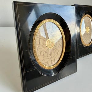 May include: Two square black picture frames with an oval centre opening. Each frame features a gold-toned decorative inner border and a textured beige insert with a circular gold seal in the centre. The black surface has thin gold lines.