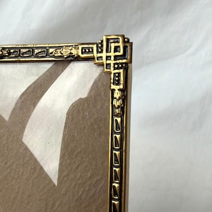 May include: Close-up of a gold-colored picture frame with intricate Art Deco-style detailing. The frame has a rectangular shape with a decorative border and corner accents. The frame is empty, with a clear glass or plastic insert.