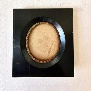 May include: A square, black picture frame with a circular opening. The frame has a decorative, gold-colored inner ring. The center of the frame is a light tan color.