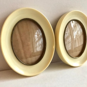 Puede incluir: Dos marcos de fotos ovalados con borde crema y marco interior bronce. Los marcos están uno al lado del otro, mostrando una estética vintage. El cristal refleja el entorno.