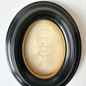 May include: Oval black picture frame with a gold inner border. The frame holds a sepia-toned portrait of a man in a suit and tie. The frame is set against a white background.