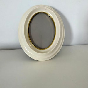 May include: Cream-colored oval picture frame with a gold-colored inner rim. The frame has a layered design and a gray interior. The frame is sitting on a white surface.