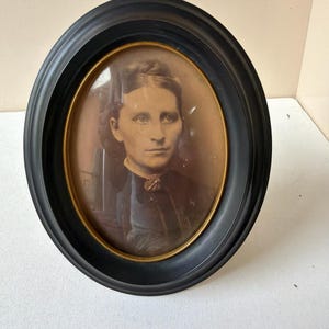May include: Oval portrait of a woman in a black frame. The photograph is sepia-toned, showing a woman with dark hair and a serious expression. The frame is black with a gold inner edge, and the portrait is set against a neutral background.