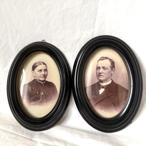 Puede incluir: Dos retratos ovalados antiguos en marcos negros. Un retrato presenta a una mujer con un vestido oscuro, el otro a un hombre con traje y pajarita. Los retratos son en tonos sepia, con una estética vintage.