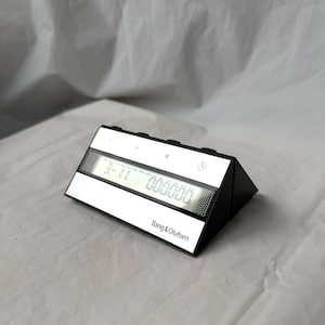 Può includere: Un orologio digitale in bianco e nero con la scritta "Bang & Olufsen" sul davanti. L'orologio mostra l'ora e ha dei pulsanti sulla parte superiore. L'orologio ha una forma triangolare.