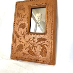 May include: A rectangular wooden picture frame with intricate floral carvings. The frame holds a black and white portrait of a man. The frame is a warm brown color and has a detailed, carved design around the edges and corners.