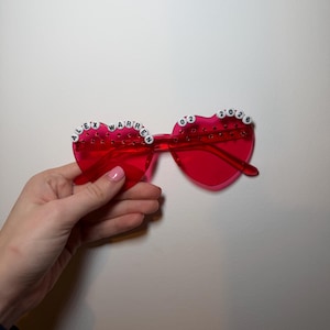 Könnte beinhalten: Rote herzförmige Sonnenbrille mit dem Text "ALEX WARREN 02 2026" in weißen Perlen auf der Oberseite. Die Sonnenbrille wird von einer Hand vor einem weißen Hintergrund gehalten.