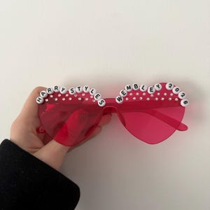 May include: Red heart-shaped sunglasses with the text "HARRY STYLES WEMBLEY 2026" spelled out in white letter beads. The sunglasses have clear rhinestones along the top edge.