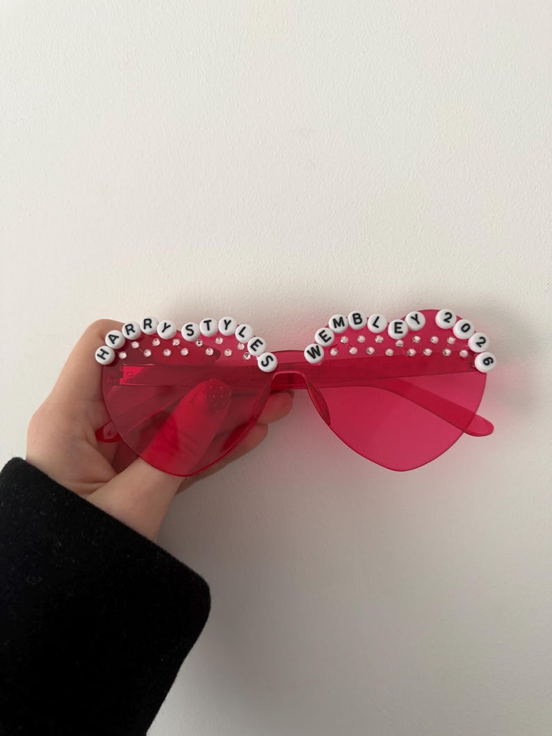 May include: Red heart-shaped sunglasses with the text "HARRY STYLES WEMBLEY 2026" spelled out in white letter beads. The sunglasses have clear rhinestones along the top edge.