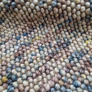 Op de afbeelding: Close-up van een getextureerd tapijt met een patroon van kleine, afgeronde, veelkleurige elementen. De kleuren zijn beige, bruin, blauw en geel, wat een natuurlijke, aardse esthetiek creëert. De tekst "Rug Haus Store" is zichtbaar.