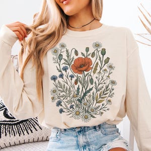May include: A cream-colored long-sleeve sweatshirt featuring a central graphic print of a wildflower bouquet. The illustration includes a large orange poppy, blue cornflowers, and white daisies with green stems and leaves arranged in a cluster.