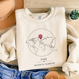 Puede incluir: Sudadera color crema con un gráfico de mapa del mundo dentro de un círculo, con un pin rojo en forma de corazón. El nombre "Emily" y las coordenadas están impresas debajo. La sudadera está doblada sobre una superficie de madera.