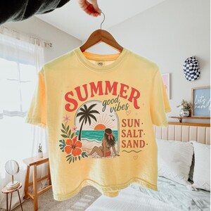 May include: A pale yellow short-sleeve t-shirt featuring a graphic print of a beach scene with a palm tree, a person sitting on the sand, and a large orange hibiscus flower. The text reads SUMMER good vibes SUN SALT SAND in red and black.