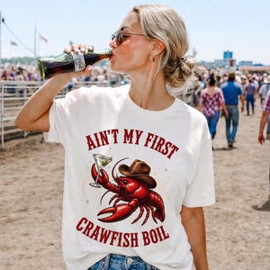 Puede incluir: Una camiseta blanca de manga corta con el gráfico de un cangrejo de río rojo que lleva un sombrero de vaquero marrón y sostiene una copa de martini. El texto "AIN'T MY FIRST CRAWFISH BOIL" está impreso en rojo sobre la ilustración.