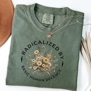 Könnte beinhalten: Olivgrünes T-Shirt mit dem Text "RADICALIZED BY BASIC HUMAN DECENCY" in einem kreisförmigen Design um einen Blumenstrauß. Eine goldene Halskette mit Anhänger liegt auf dem Shirt. Ein brauner Hut ist teilweise in der oberen linken Ecke zu sehen.