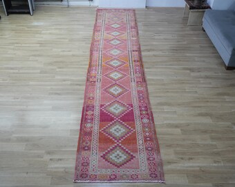 May include: A long, rectangular rug with a pink and orange diamond pattern. The rug has a central design of repeating diamond shapes in shades of pink, orange, and white, framed by a border with floral and geometric motifs. The rug is placed on a light wood floor.