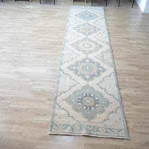 May include: A long, narrow runner rug featuring a repeating medallion pattern in muted shades of sage green, beige, and soft blue. The design consists of symmetrical floral-inspired motifs centered within geometric borders on a cream-colored background.