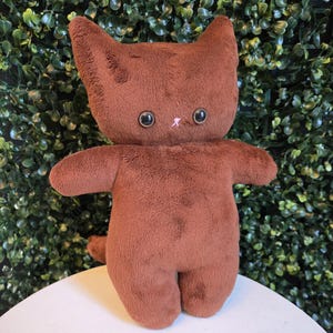 May include: A brown plush cat doll with large, round black eyes and a small pink nose. The cat has a soft, textured appearance and is standing upright with its arms outstretched. The background is a green hedge.