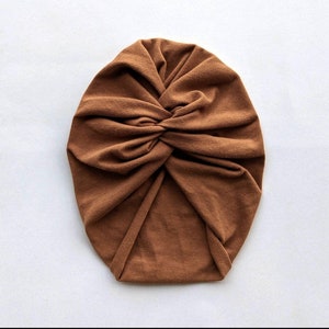 May include: A brown turban hat made from soft fabric. The hat has a twisted knot detail at the front, offering a stylish and comfortable head covering. The hat is a solid colour and is suitable for various occasions.