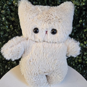 May include: A cream-coloured, cat-shaped soft toy with large black button eyes and a small red embroidered nose. The toy has a fluffy texture and a simple design, suitable for children or collectors.