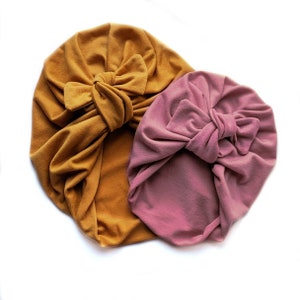 May include: Two baby turbans. One is mustard yellow, the other is dusty rose. Both have a large bow on the front. The fabric appears to be soft and comfortable. The turbans are likely for infants or toddlers.