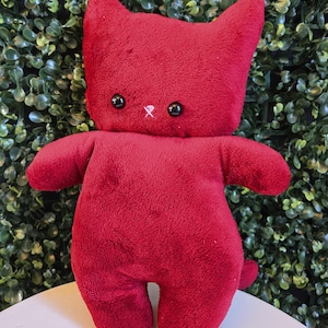 May include: A red plush cat doll with black button eyes and a small white cross stitched below the eyes. The cat has a small tail and is standing on a white surface in front of a green leafy background.