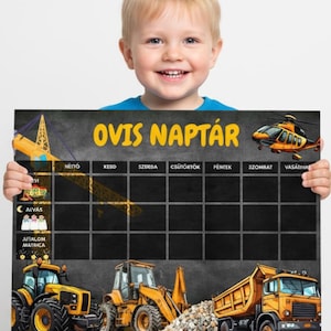 Puede incluir: Un niño sostiene un calendario negro y amarillo con temática de construcción con el texto "OVIS NAPTÁR" en amarillo. El calendario presenta ilustraciones de vehículos de construcción, incluyendo un tractor, una excavadora y un camión volquete, y un helicóptero.