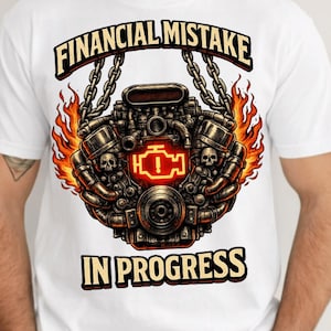 May include: White t-shirt with a detailed engine graphic, flames, and chains. The text "FINANCIAL MISTAKE" is above the engine, with "IN PROGRESS" below. The engine graphic includes a glowing check engine light.