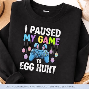 Könnte beinhalten: Schwarzes Sweatshirt mit dem Text "I PAUSED MY GAME TO EGG HUNT". Das Design zeigt einen blauen Gamecontroller mit Hasenohren und hängenden Ostereiern in Pastellfarben. Ein lustiges, festliches Design für Ostern.
