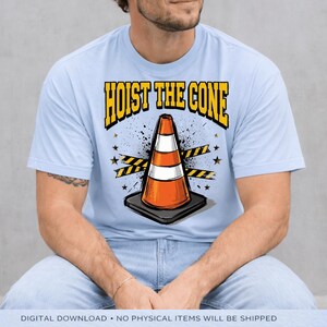 Puede incluir: Camiseta azul claro con el texto "HOIST THE CONE" en amarillo y negro. El gr&aacute;fico presenta un cono de tr&aacute;fico naranja y blanco sobre una base negra, con estrellas y cintas de precauci&oacute;n. Descarga digital.